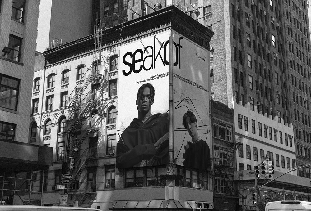 Black and white photo of a city block featuring a large billboard on a building corner. The billboard displays the word "Seekout" and images of two individuals. Surrounding buildings and urban structures are visible in the background.