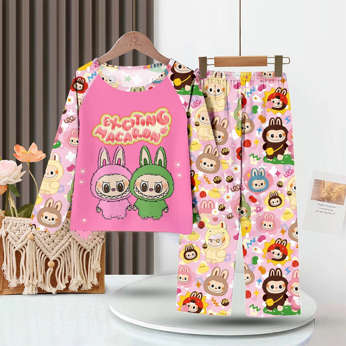 The LABUBU Baby Pajama Set by LABUBU is a cozy, milk silk kids sleepwear ensemble featuring a colorful design with cartoon bunnies, a pink top reading "Exciting Vacation," and pants with a macaron pattern, enhanced by charming macrame decor and a potted plant in the background.