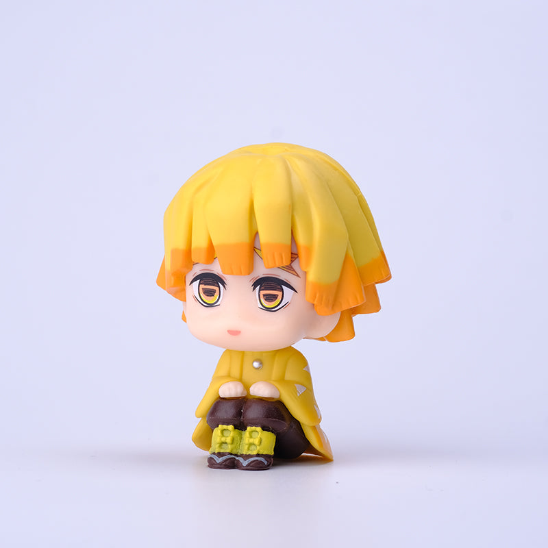 The Demon Slayer Figure Set features an anime-style figure with yellow hair and an orange-yellow coat, hugging its knees on a white background. Ideal for any fan, this 7cm PVC figurine from the Demon Slayer: Kimetsu no Yaiba Collection has a neutral expression and expressive eyes.