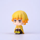The Demon Slayer Figure Set features an anime-style figure with yellow hair and an orange-yellow coat, hugging its knees on a white background. Ideal for any fan, this 7cm PVC figurine from the Demon Slayer: Kimetsu no Yaiba Collection has a neutral expression and expressive eyes.
