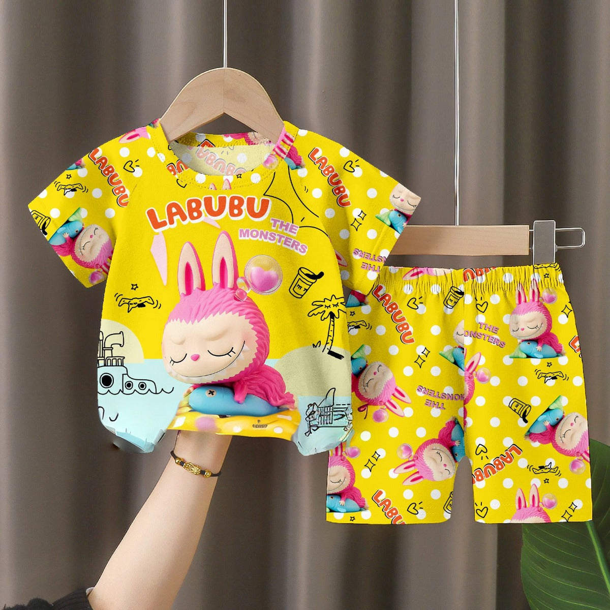 The LABUBU Baby Pajama Set in bright yellow milk silk features a playful "The Monsters" theme with cartoon-style illustrations of a pink-haired character, pink bunny, and colorful patterns, all adorned with the word "LABUBU.