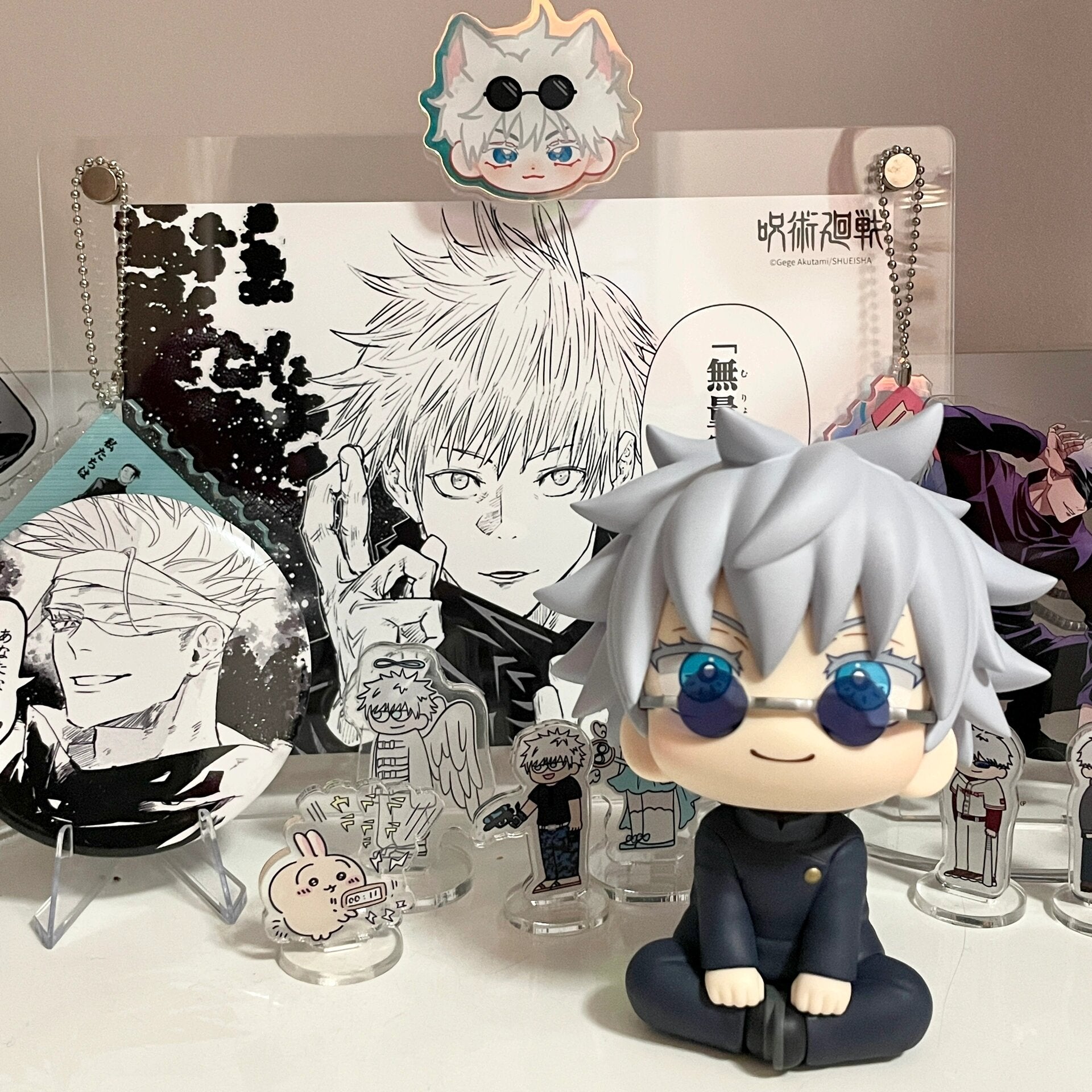 A display of anime items features the Jujutsu Kaisen Gojo Satoru 12cm Rotatable Head Figure with gray hair and glasses, surrounded by character cutouts and manga pages, making it a must-have collectible for fans.