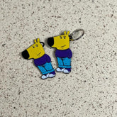 Two identical acrylic keychains from the "Chill Guy Keychain Set – 10-Piece Meme-Inspired Collection" feature a yellow, dog-like character with a purple shirt, blue pants, and white shoes on a speckled beige surface.