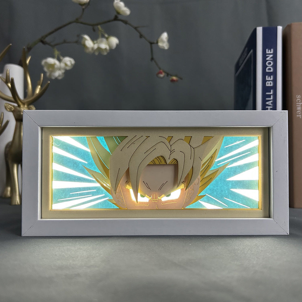 The Seakoff Dragon Ball Z Anime Light Box showcases Super Saiyan Goku in 3D with spiky hair and glowing eyes against a vibrant background. It’s encased in a white frame on a table next to gold deer figurine, books, and white flowers, offering a 16-color adjustable LED light display.