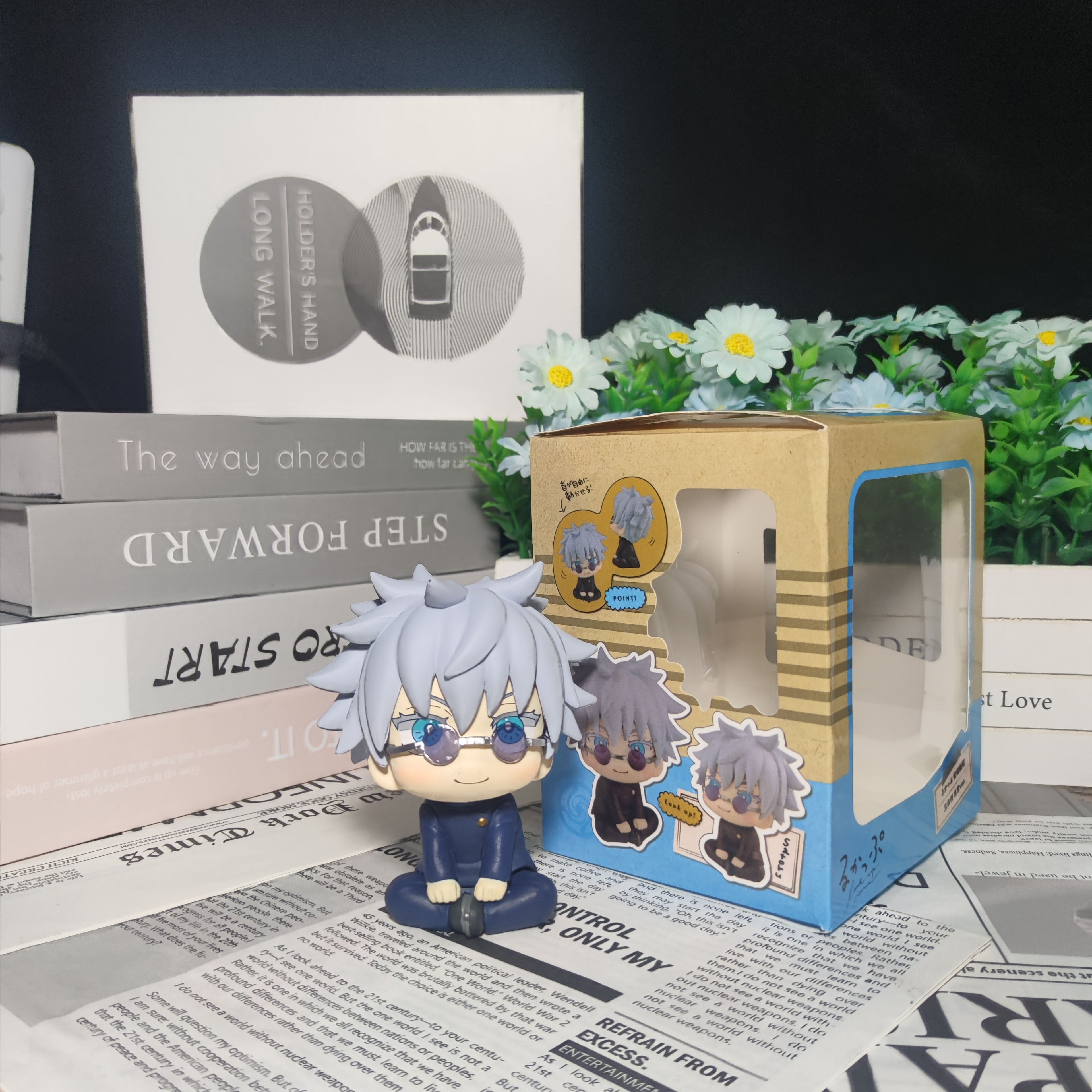 The Gojo Satoru Anime Figure by Jujutsu Kaisen, a 12cm chibi-style collectible with gray hair and glasses, sits beside a themed box. It’s set elegantly against stacked books and a small plant with white flowers on a reflective surface.