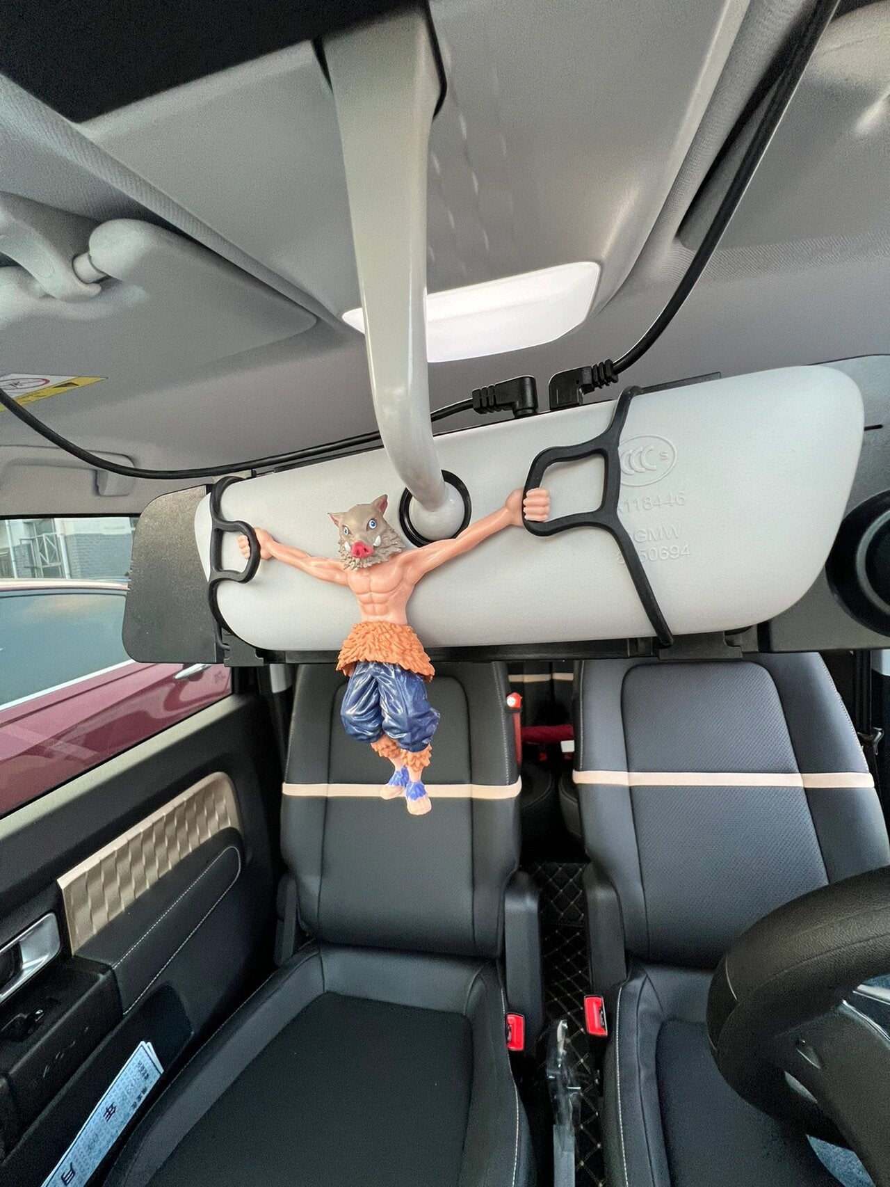 An Inosuke Hashibira Hanging Figure from Demon Slayer, featuring exaggerated muscles, humorously appears to hold up a large gray foam roller under a car&
