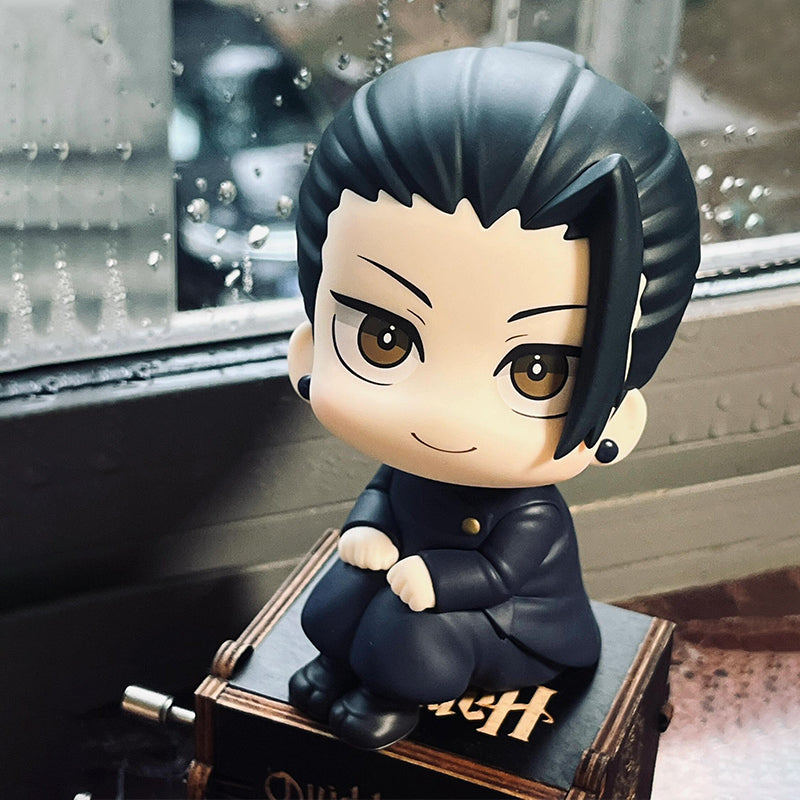 A chibi-style Suguru Geto figure from the Jujutsu Kaisen collection features a rotatable head and is depicted sitting cross-legged with black hair and earrings, in a black outfit, on a small wooden crate by a rain-dappled window, sporting a slight smile.