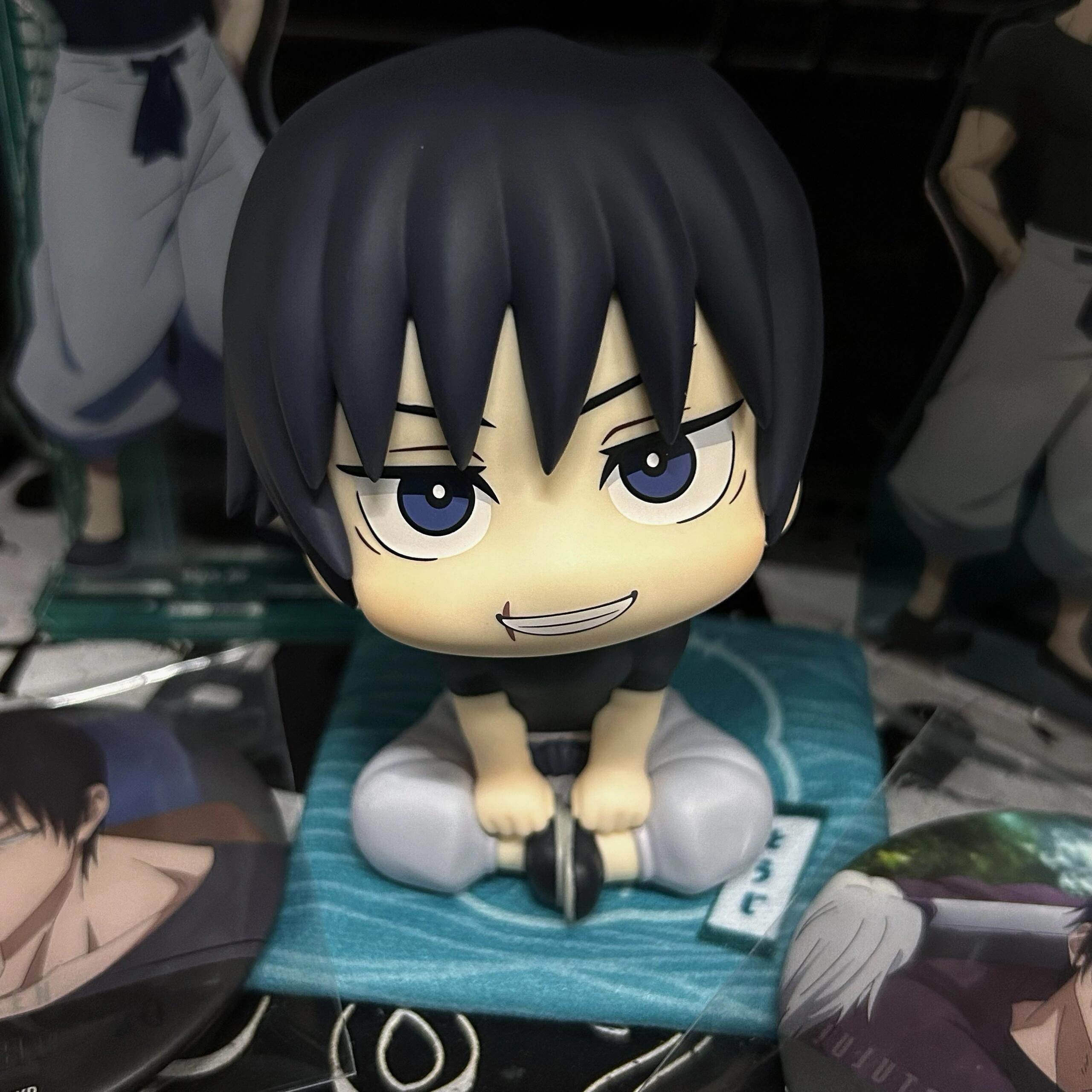 A 12cm Toji Fushiguro figure with black hair and blue eyes, part of the Jujutsu Kaisen collection, sits cross-legged on a small cushion among anime collectibles like circular badges and character stands. This rotatable head action figure is a must-have collectible for fans.