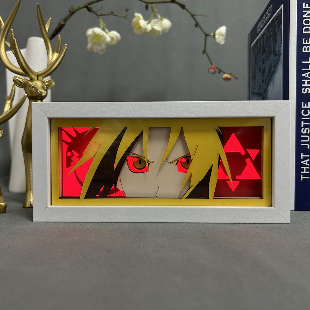 A framed artwork displays an anime character with intense eyes set against a red triangle backdrop, hinting at alchemy symbols. The white frame sits alongside decorative pieces, including a Fullmetal Alchemist LED Light Box – 3D Edward Elric Anime Lamp, a golden antler sculpture, and a book.