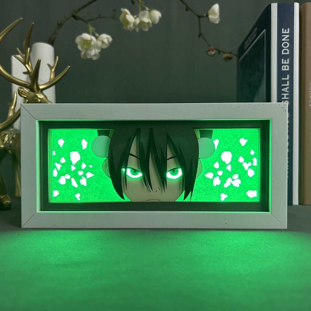 The Toph Beifong LED Light Box, branded by Avatar the Last Airbender, showcases an anime-style face with dark hair and glowing eyes in green. The character stands out against abstract green and white designs, while books and a decorative antler are partially visible.