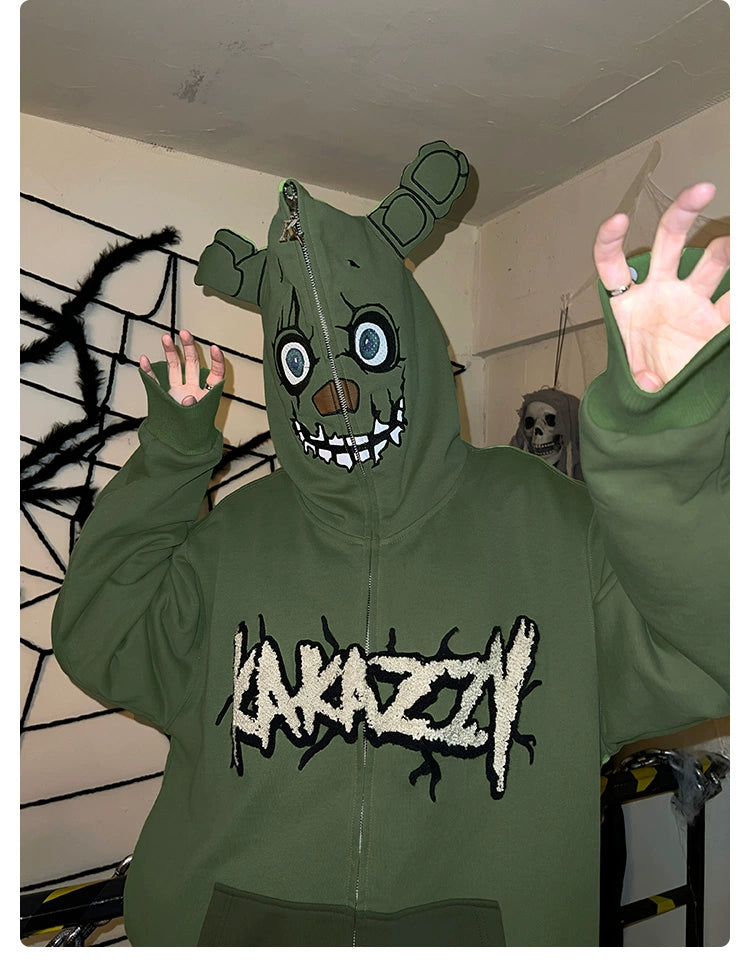 A person wears a Seakoff Springtrap-Inspired Hoodie, a green oversized zip-up with cartoon face and bear ears. Amid Halloween skeletons and spider webs, it echoes the creepy vibe of Five Nights at Freddy&