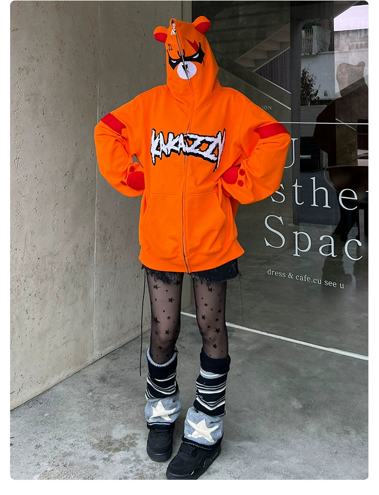 A person wears a Seakoff Furious Orange Bear Hoodie with cartoon ears and an Angry Bear Face graphic, paired with star-patterned leggings, shin-high leg warmers, and sleek black shoes against a backdrop of a concrete wall and glass window.