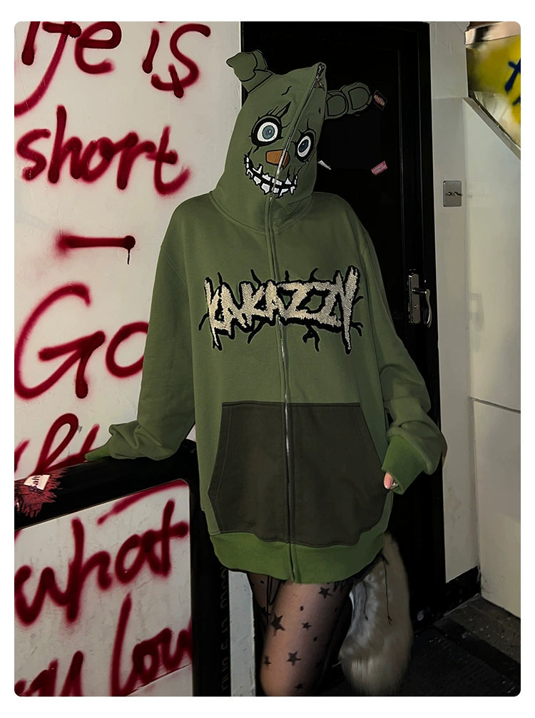 A person stands indoors wearing a Seakoff Springtrap-Inspired Hoodie, a green 400GSM oversized zip-up with a mischievous cartoon face hood, perfect for Five Nights at Freddy&