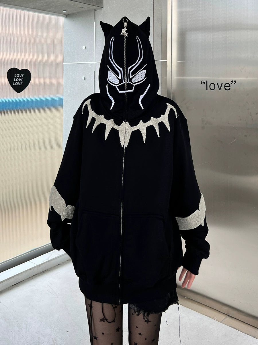 A person wearing a Seakoff Black Panther-Inspired Hoodie stands in a metallic-walled room. The oversized zip-up sweatshirt, loved by streetwear fans, features cat ears, a stylized face design with a jagged white chest pattern. The word "love" and heart-shaped decor adorn the wall.