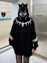 A person wearing a Seakoff Black Panther-Inspired Hoodie stands in a metallic-walled room. The oversized zip-up sweatshirt, loved by streetwear fans, features cat ears, a stylized face design with a jagged white chest pattern. The word "love" and heart-shaped decor adorn the wall.