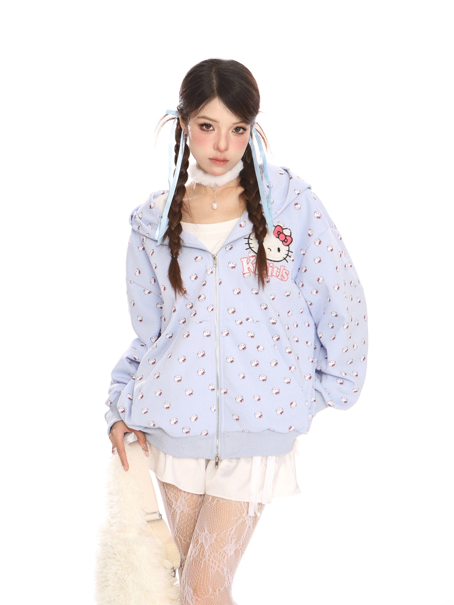 A woman with braided hair wears a Hello Kitty Pink Kawaii Hoodie by Seakoff, featuring an oversized zip-up with cat ear hood and all-over print. She pairs it with a white skirt, lace stockings, and a choker against a crisp white background.