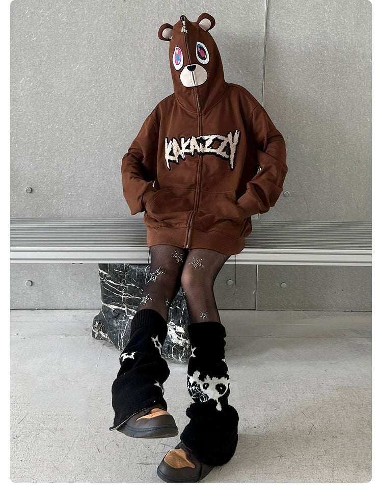 Wearing the Seakoff Cute Bear Hoodie—an oversized 400GSM brown zip-up streetwear sweatshirt with ears—a person sits on a bench. The hoodie features a bear face on the hood, paired with black and white leg warmers and black shoes, making them stand out against a plain gray wall.