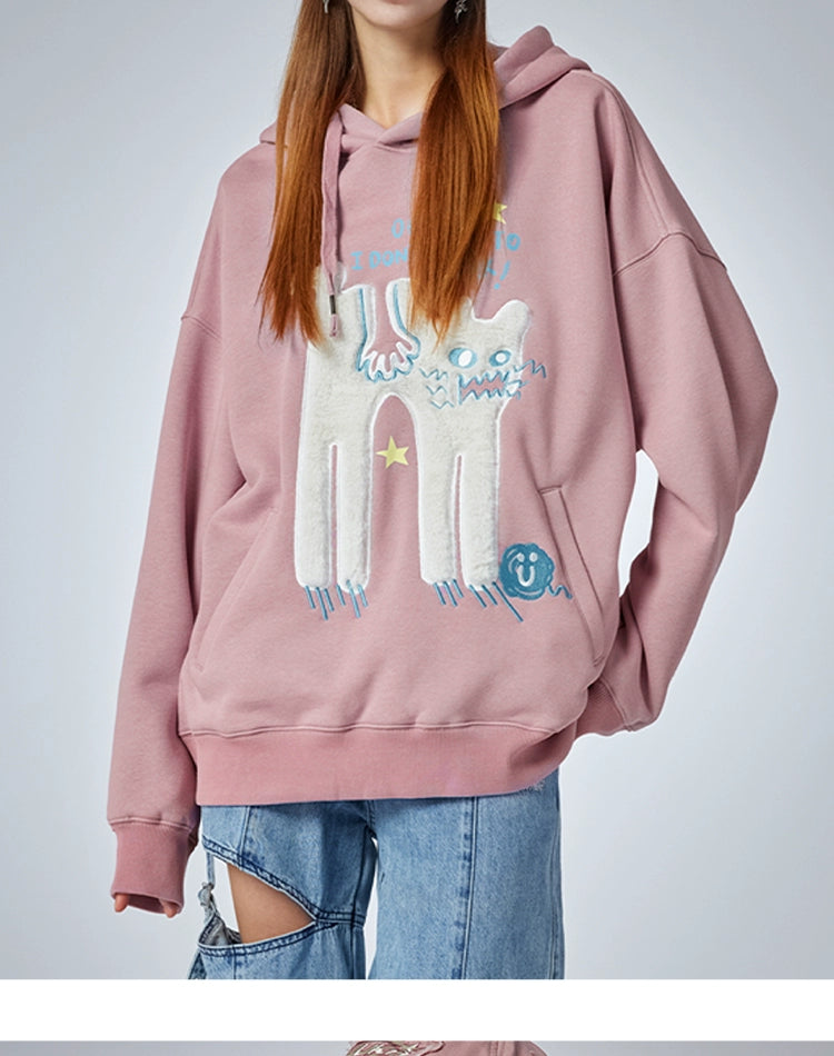A person wears the Seakoff "Oh No! I Don’t Want to Work!" oversized pink hoodie, featuring a cute lazy cat design. The relaxed fit and long sleeves pair perfectly with ripped blue jeans against a plain, light background, embodying casual streetwear vibes.