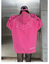 A person stands with their back to the camera, wearing a Seakoff Spider-Inspired Hoodie—an oversized 400GSM pink zip-up sweatshirt ideal for superhero fans. It sports a white spiderweb design and is paired with a matching pink cap and fishnet leggings. The backdrop is a metallic wall with partial text visible.