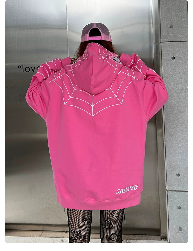 A person stands with their back to the camera, wearing a Seakoff Spider-Inspired Hoodie—an oversized 400GSM pink zip-up sweatshirt ideal for superhero fans. It sports a white spiderweb design and is paired with a matching pink cap and fishnet leggings. The backdrop is a metallic wall with partial text visible.