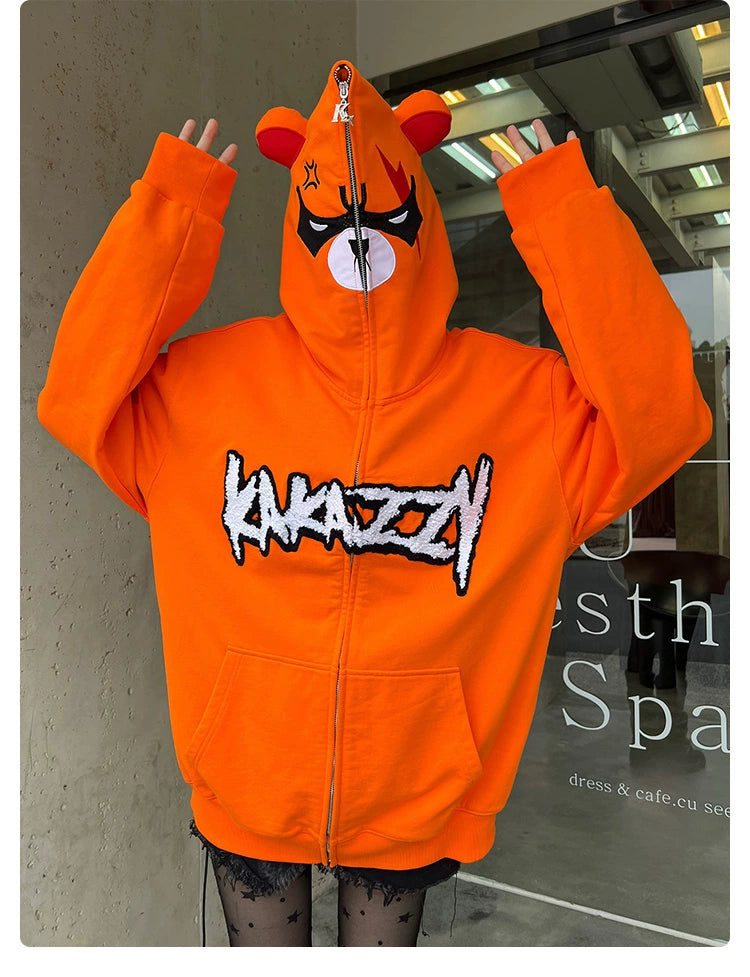 A person in front of a glass-doored storefront sports the Seakoff Furious Orange Bear Hoodie, made from premium 400GSM cotton. It features an angry bear face with ears, partially zipped over their face, vibrant orange with stylized eye mask graphics and red "ANARCHY" embroidery.