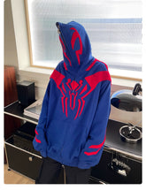 The person wears a Seakoff Superhero Spider Hoodie, featuring a blue and red emblemed full zip-up face cover, embodying Spider-Man. They are in a modern room with sleek audio equipment and a stylish lamp in the background.