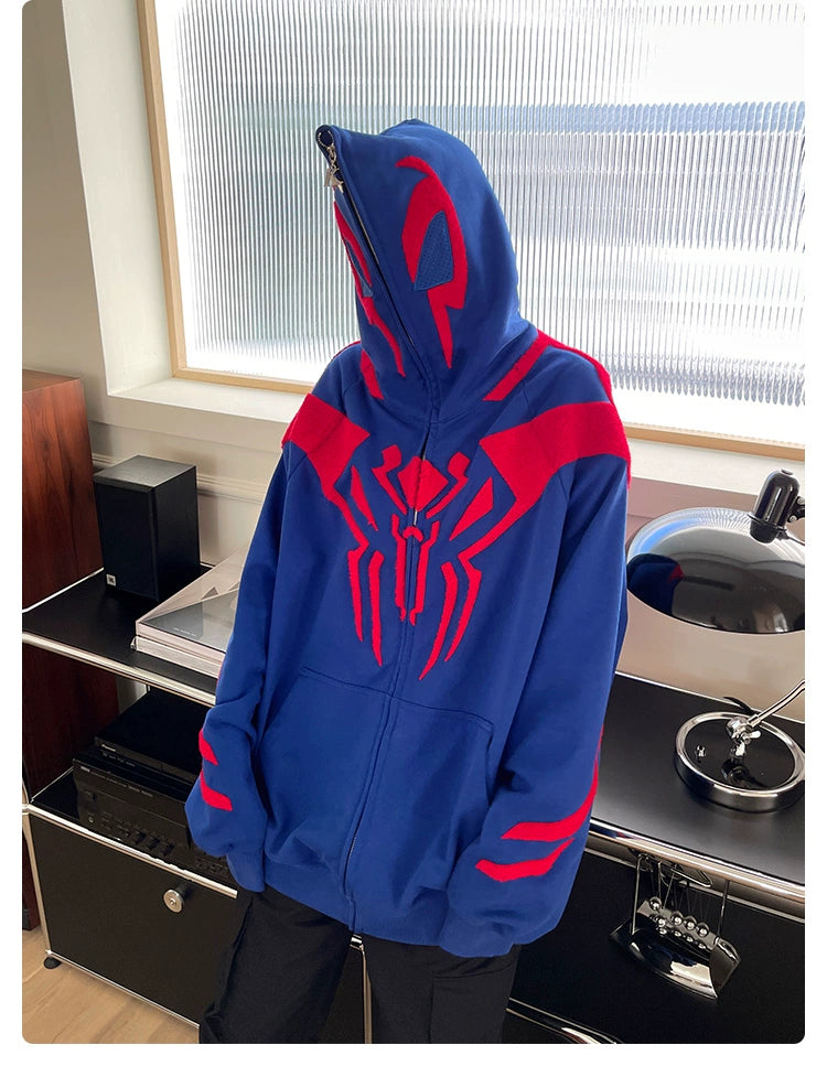 The person wears a Seakoff Superhero Spider Hoodie, featuring a blue and red emblemed full zip-up face cover, embodying Spider-Man. They are in a modern room with sleek audio equipment and a stylish lamp in the background.