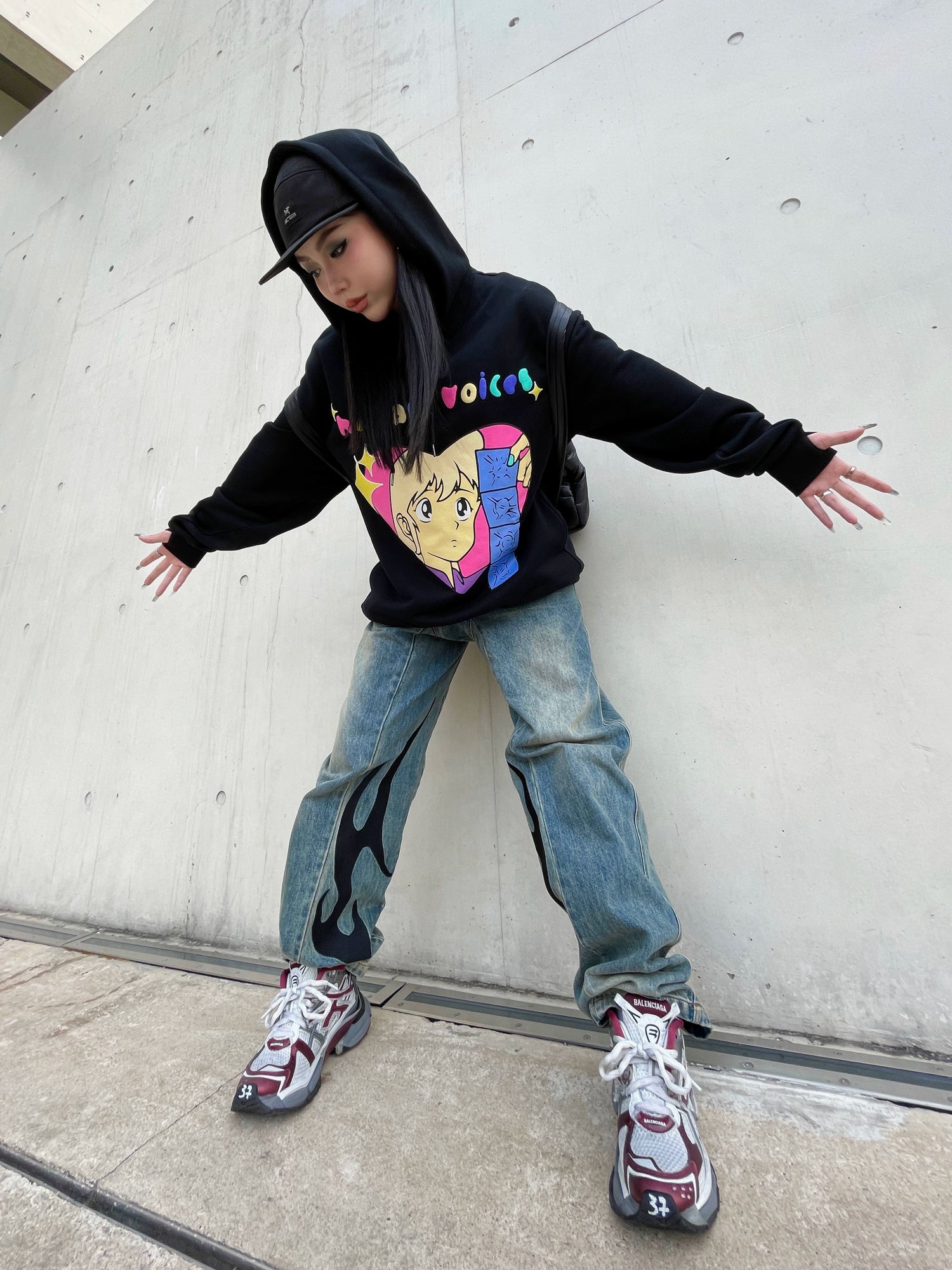 A person in a black Seakoff Retro Anime Hoodie with a Human Voices graphic and drop shoulder design stands against a concrete wall wearing jeans with flame designs on one leg, a black cap, and sneakers featuring maroon accents, arms outstretched evoking nostalgic vibes.
