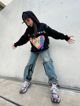 A person in a black Seakoff Retro Anime Hoodie with a Human Voices graphic and drop shoulder design stands against a concrete wall wearing jeans with flame designs on one leg, a black cap, and sneakers featuring maroon accents, arms outstretched evoking nostalgic vibes.