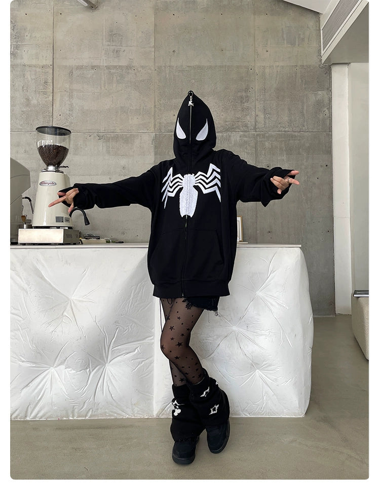 A person in a Seakoff Venom Spider Hoodie - 400GSM Oversized Black and White Zip-Up Sweatshirt stands with outstretched arms, wearing patterned tights and black boots, set against a white couch and coffee grinder backdrop, perfectly capturing the edgy streetwear vibe fans adore.