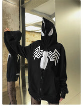 A person wearing the Seakoff Venom Spider Hoodie, a 400GSM oversized black and white zip-up sweatshirt with a white spider emblem and eye design, stands in front of a mirror. The hoodie zips up to cover the face and pairs with patterned tights against an industrial-style background.