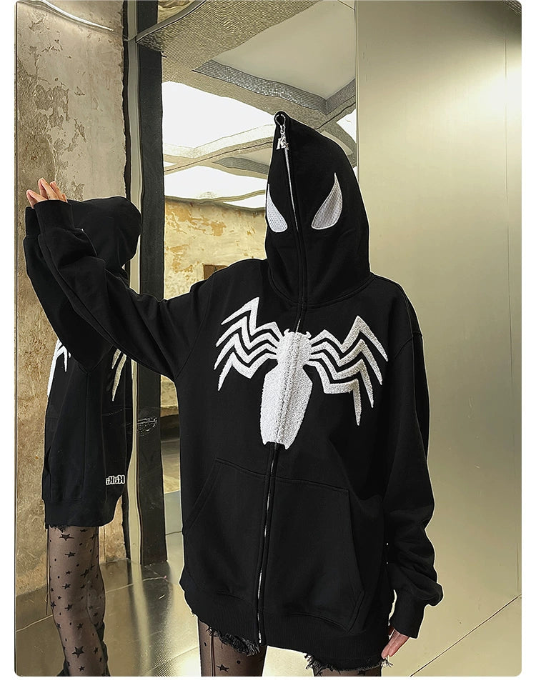 A person wearing the Seakoff Venom Spider Hoodie, a 400GSM oversized black and white zip-up sweatshirt with a white spider emblem and eye design, stands in front of a mirror. The hoodie zips up to cover the face and pairs with patterned tights against an industrial-style background.