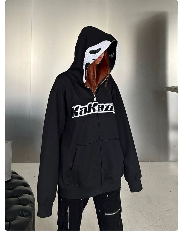 A person wears a Seakoff "Scream-Inspired Hoodie," an oversized black zip-up sweatshirt with a Ghostface design. Their long hair falls over the hood while they stand in a minimalistic room featuring a sleek silver wall and stylish black cushioned seat—ideal for horror fans.
