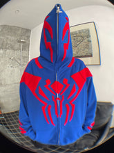 A person in a Seakoff Superhero Spider Hoodie, an oversized 400GSM blue sweatshirt with a red spider emblem, has their face obscured by the fully zipped hood. They stand indoors against a gray textured wall and framed artwork, making a bold streetwear statement.