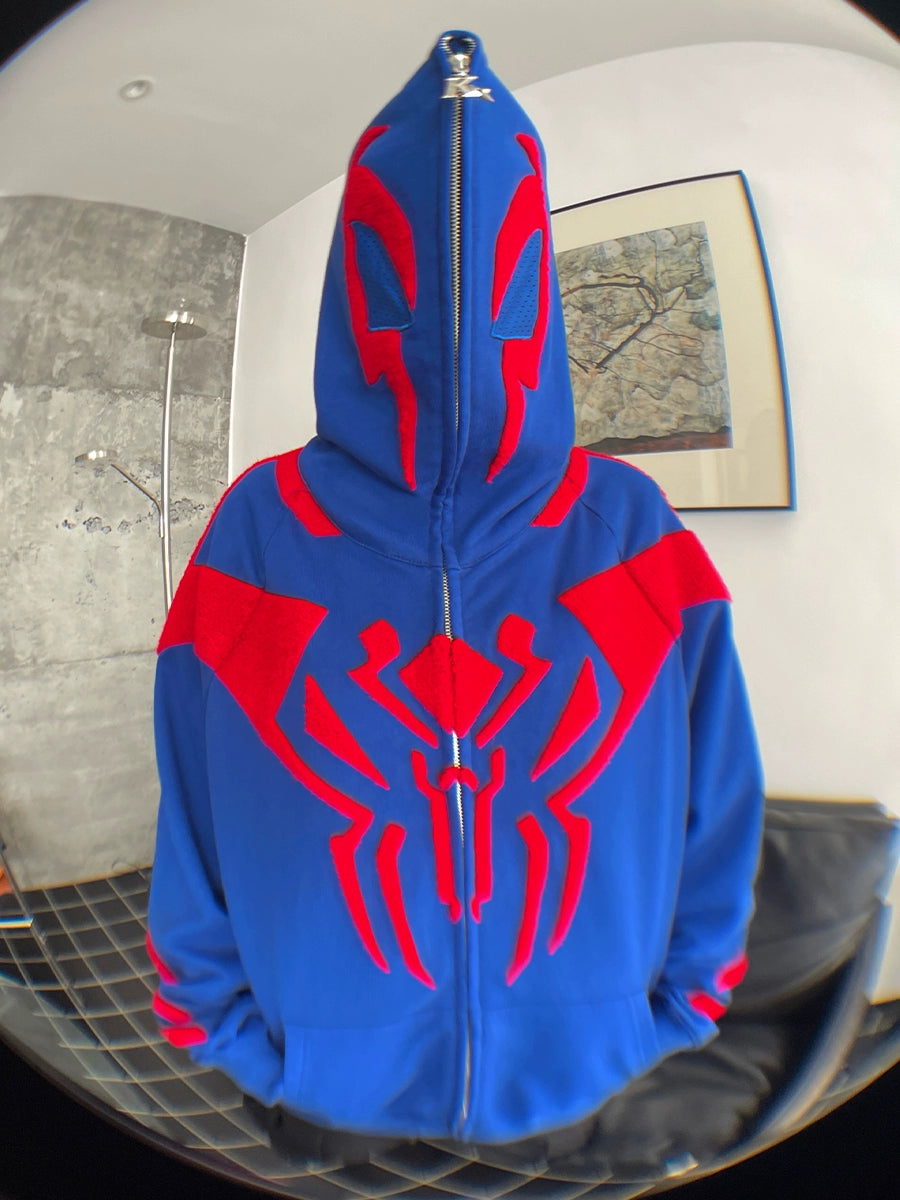 A person in a Seakoff Superhero Spider Hoodie, an oversized 400GSM blue sweatshirt with a red spider emblem, has their face obscured by the fully zipped hood. They stand indoors against a gray textured wall and framed artwork, making a bold streetwear statement.