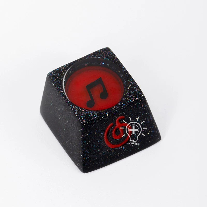 A black keycap with a shimmering surface features a red circular top embossed with a musical note. Crafted from high-quality resin, its sides have speckled details and it sports "The Keycap" logo. An ideal addition to the Naruto Elements Keycap Set by Naruto—with Hidden Leaf, Akatsuki, and more.