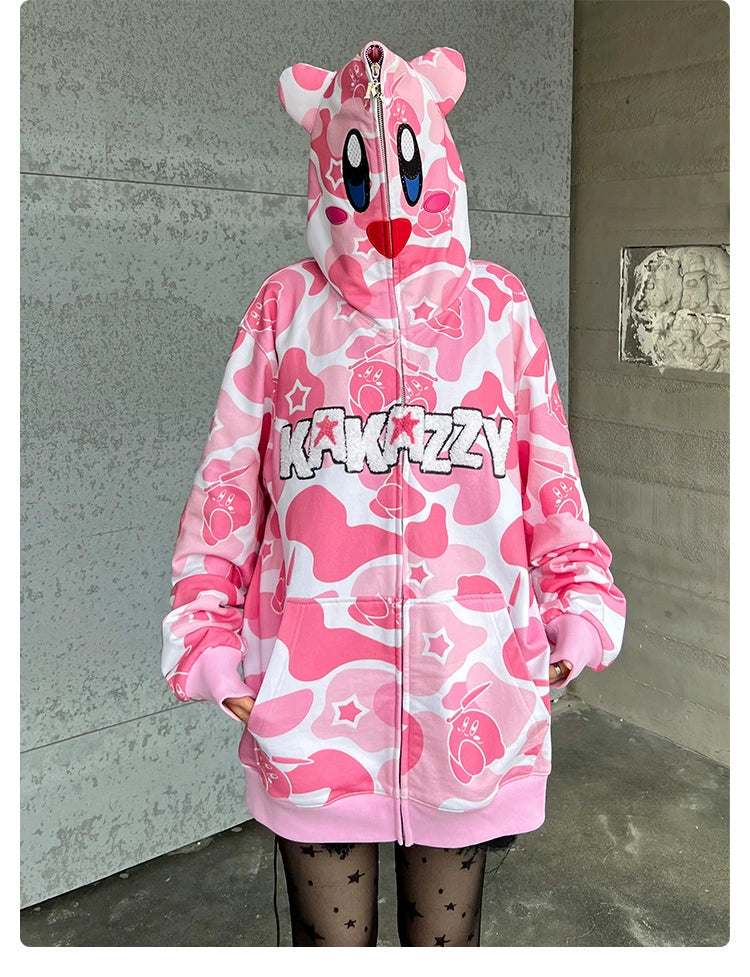 A person wears a Seakoff Pink Kirby-Inspired Hoodie featuring a trendy camo print, 3D ears, and "KAKAZZY" text, paired with star-patterned black tights for a playful look.