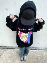 A person with their face hidden under a black cap is wearing a Seakoff Retro Anime Hoodie adorned with colorful text and a cartoon face. They are holding their long hair, complemented by jeans and sneakers, against a concrete wall backdrop, embodying nostalgic vibes.