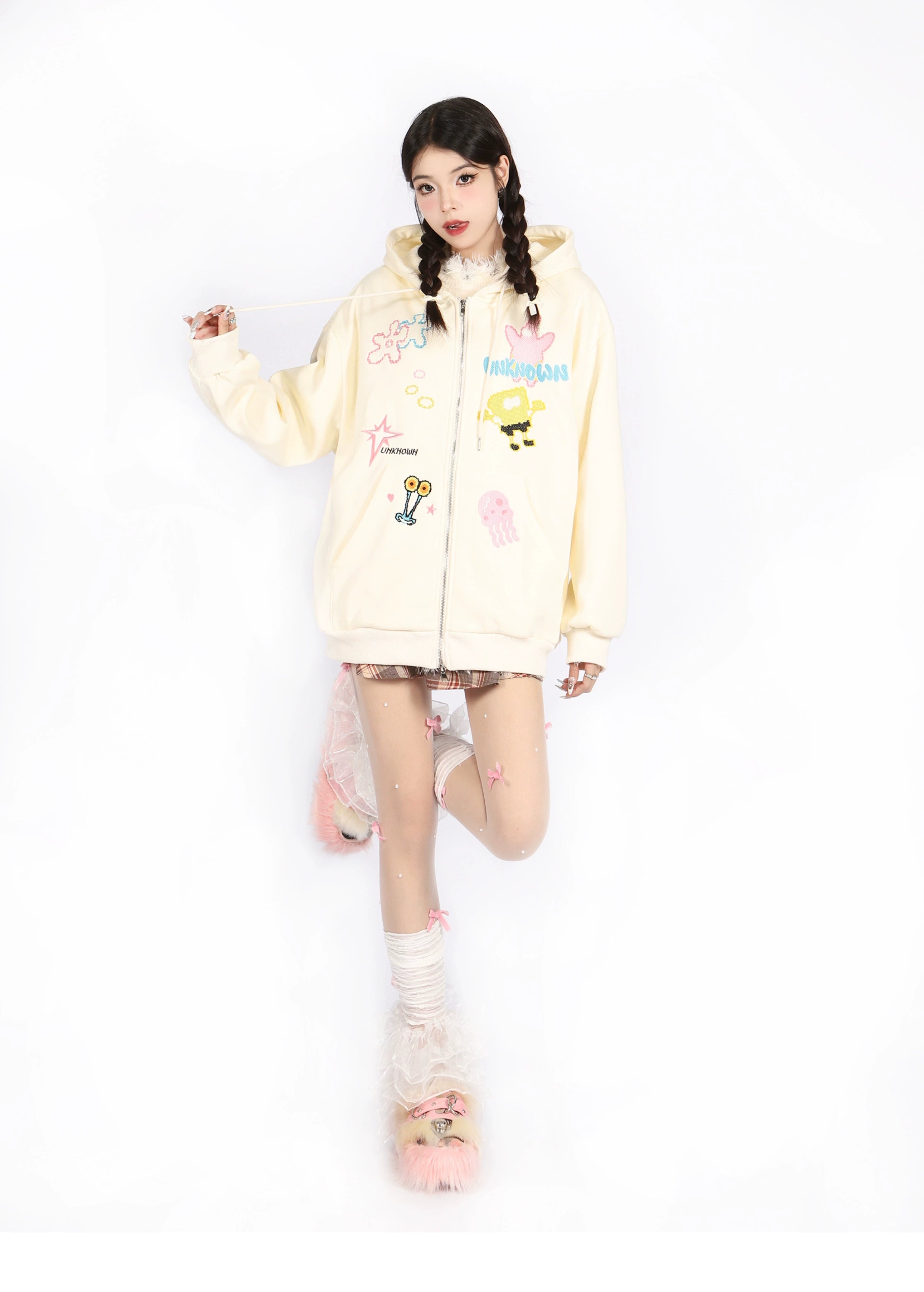 Against a white backdrop, a braided-haired individual flaunts anime fashion in a Seakoff Kawaii Pastel Cartoon Hoodie paired with a short skirt, patterned tights, and fuzzy pink slippers.