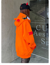 A person with long hair is wearing a Seakoff Furious Orange Bear Hoodie—crafted from premium 400GSM cotton with an angry bear design and red embroidery. They also wear a black cap with red horns and patterned tights, standing by a concrete wall adorned with signage.