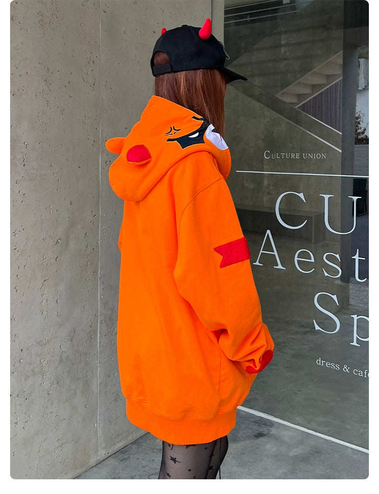 A person with long hair is wearing a Seakoff Furious Orange Bear Hoodie—crafted from premium 400GSM cotton with an angry bear design and red embroidery. They also wear a black cap with red horns and patterned tights, standing by a concrete wall adorned with signage.
