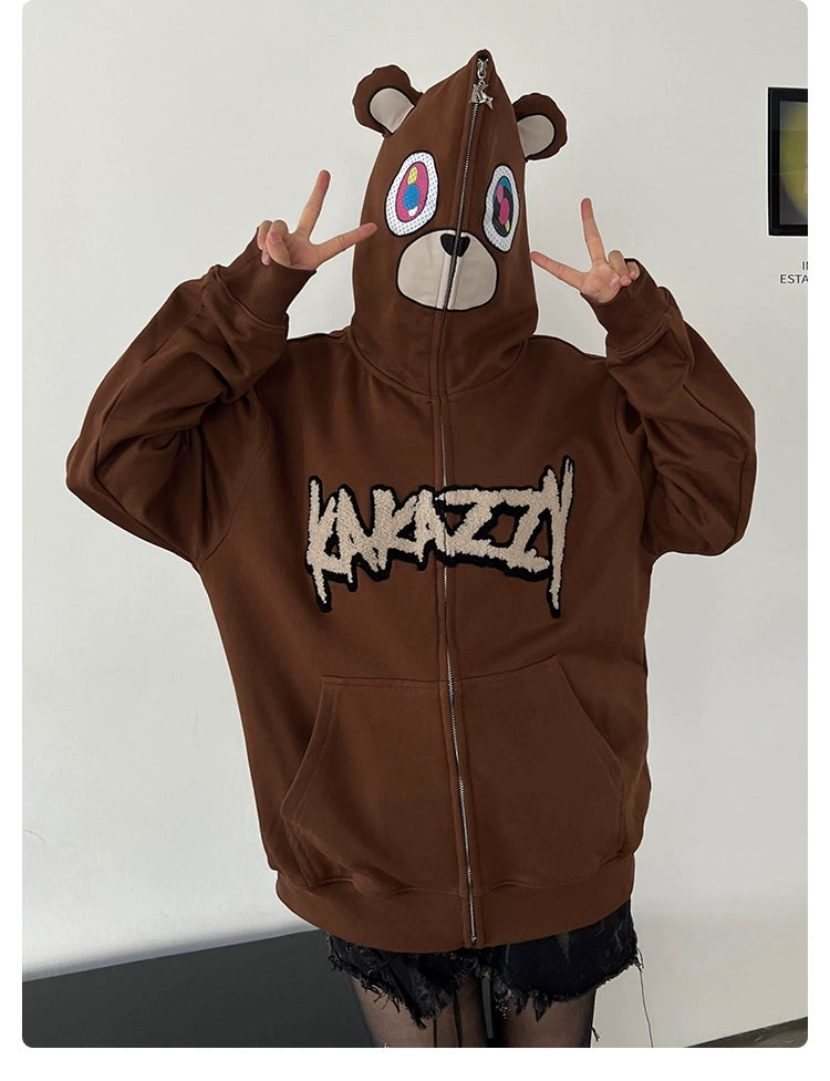 Wearing the Cute Bear Hoodie by Seakoff, an oversized 400GSM brown zip-up featuring colorful eyes and ears. "ANAZY" on the front adds flair as they strike a playful, streetwear-inspired pose with dual peace signs.