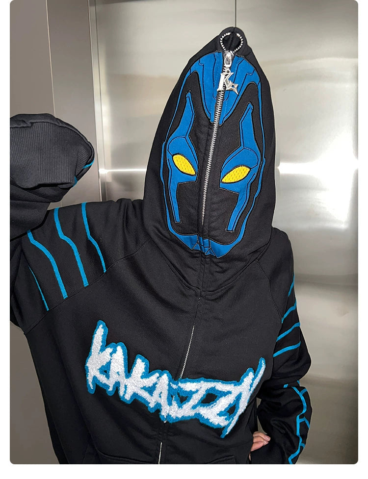 Someone is wearing the black Seakoff Cyberpunk Superhero-Inspired Hoodie. The hood, zipped completely to hide the face, features a blue and yellow mask design. "KARAZZY" in white and blue stands out on a metallic background—ideal for Sci-Fi fans with a taste for futuristic fashion.