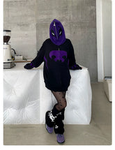 A person in a Seakoff Dark Hero Hoodie with a purple design, black star-patterned tights, and black boots stands by a white counter in a concrete-walled room. A coffee grinder sits on the counter.
