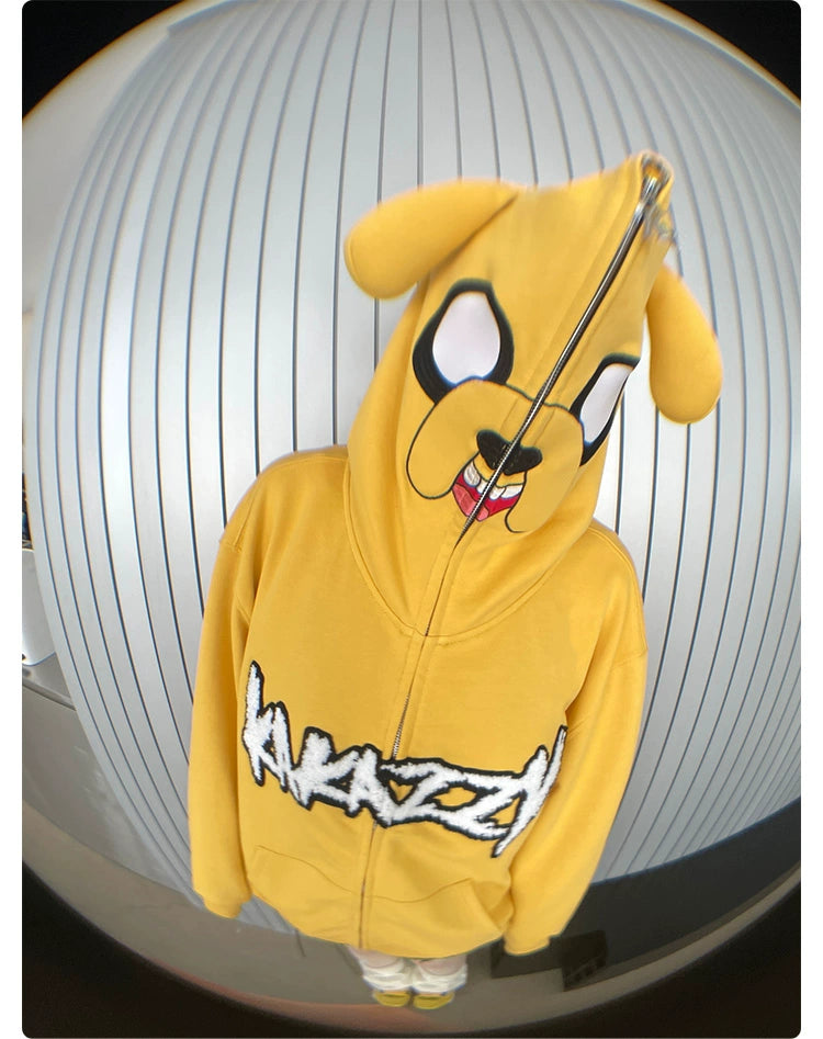A person wears the Kawaii Yellow Dog Hoodie by Seakoff, featuring a cartoon dog face with ears on the hood. The bright yellow oversized zip-up displays "KRAZZY" and makes a bold statement in anime streetwear against a striped wall.
