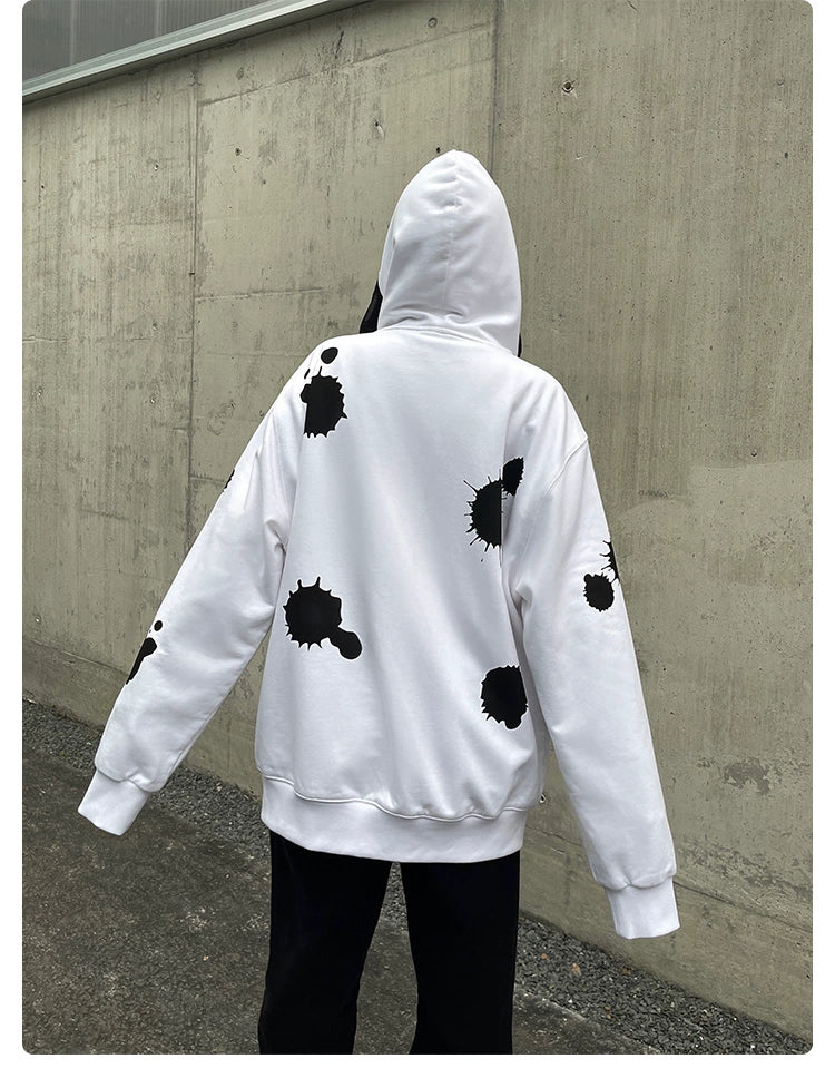 A person faces away from the camera, wearing a Seakoff zip-up hoodie named "Monochrome Ink Splatter" made from 400GSM cotton. Outdoors by a concrete wall in black pants, this outfit perfectly embodies edgy streetwear styling.