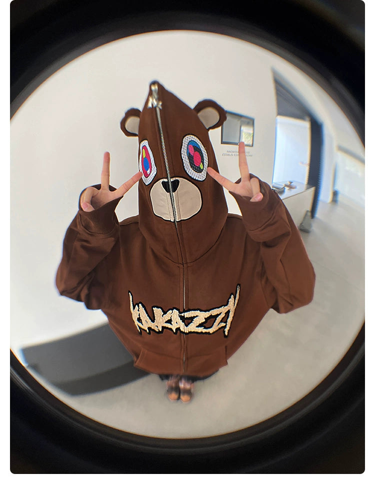 Wearing the Seakoff Cute Bear Hoodie, a 400GSM oversized brown zip-up sweatshirt with ears, an individual flashes peace signs. The playful bear face with multicolored eyes is captured creatively by a fisheye lens against a white wall and light floor, emphasizing their streetwear style.