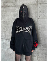 Wearing a black Berserk-Inspired Griffith Dark Knight Hoodie, zipped up to cover their face, the person stands against a concrete wall. The oversized streetwear features bold graphics and red sleeve symbols, paired with patterned tights for a Griffith-inspired look.