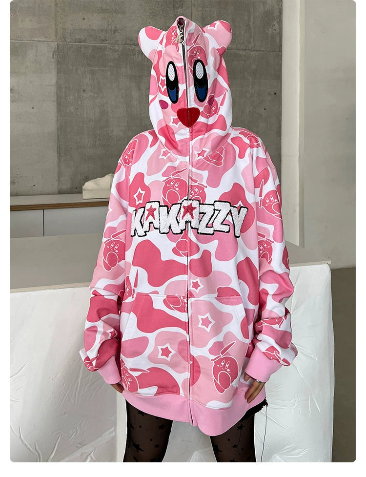 A person is wearing the Seakoff Pink Kirby-Inspired Hoodie in camo print, fully zipped to cover their face. It features cartoon eyes, a mouth, and 3D ears with "Kakazzy" on the front, set against a plain, modern room.