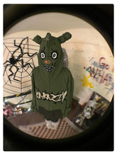 In a room decorated with wall graffiti and a large spider decoration, someone wears a Seakoff Springtrap-Inspired Hoodie. This oversized, 400GSM green zip-up sweatshirt from Five Nights at Freddy&